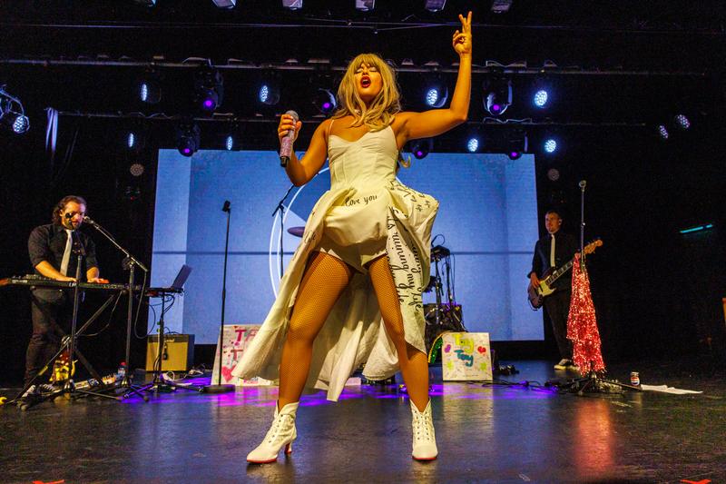 Gold dress, arms raised, singing with the band behind